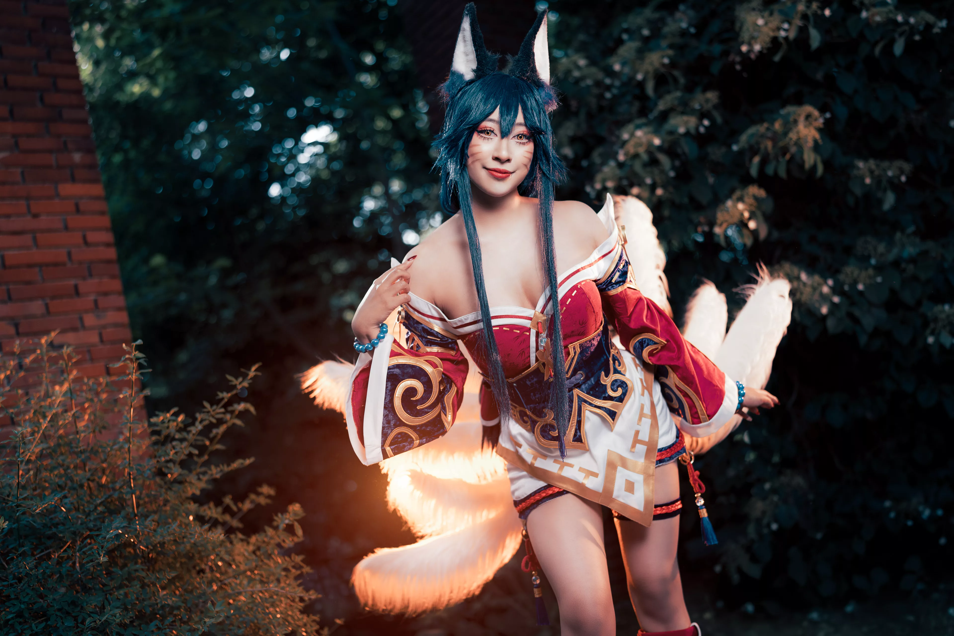 Ahri cosplay by Vivi (@vivviy.cos): nine-tailed fox in a red and blue kimono, glowing tails, in a garden at dusk